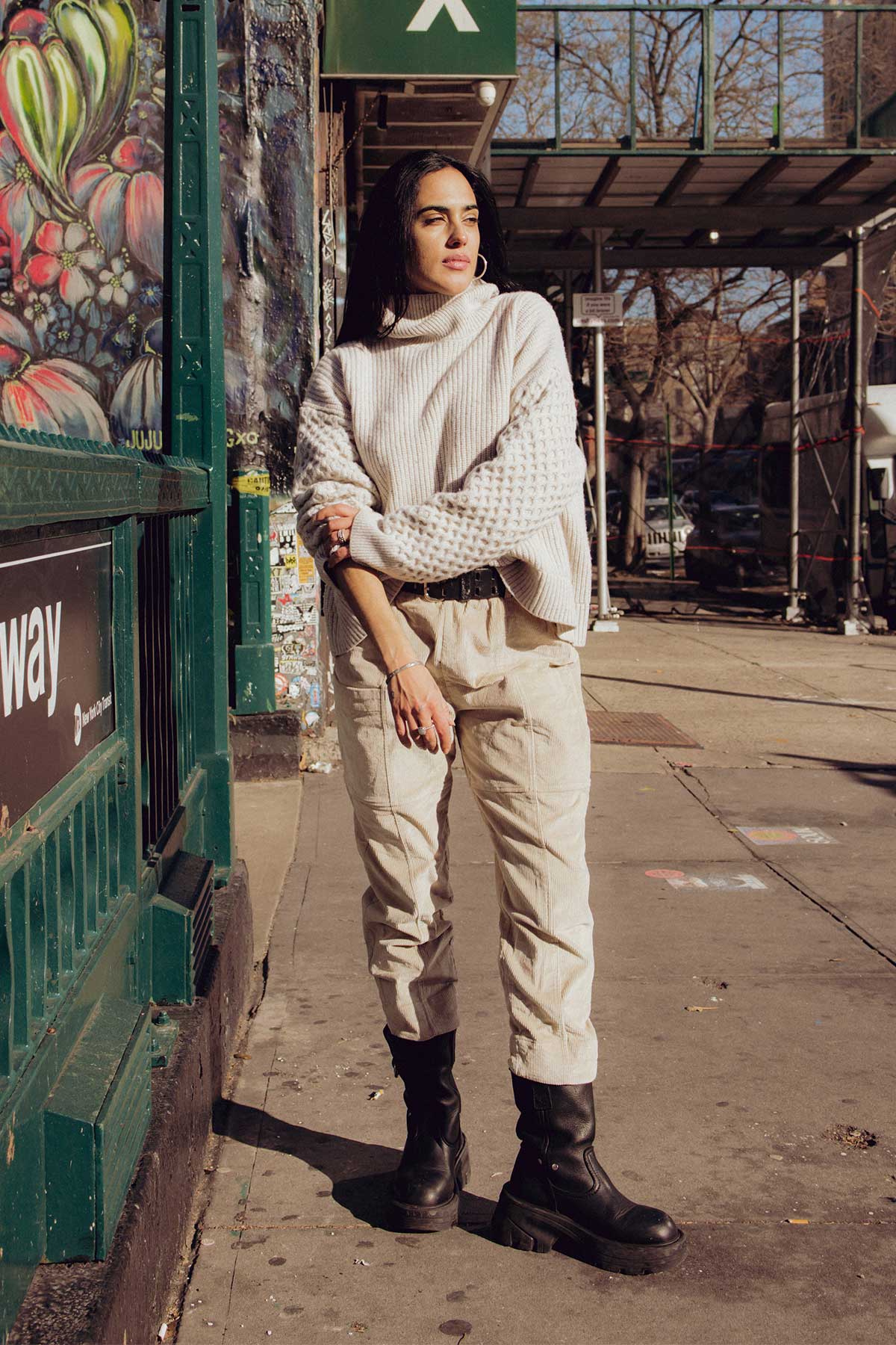 Female model is standing on a foot path wearing a textured cream knitted jersey, bone coloured corduroy tapered trousers and chunky black boots