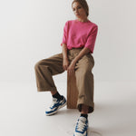 A female model sits on a wooden block in a pink t.shirt with baggy brown cuffed hem pants and sneakers against an ivory backdrop