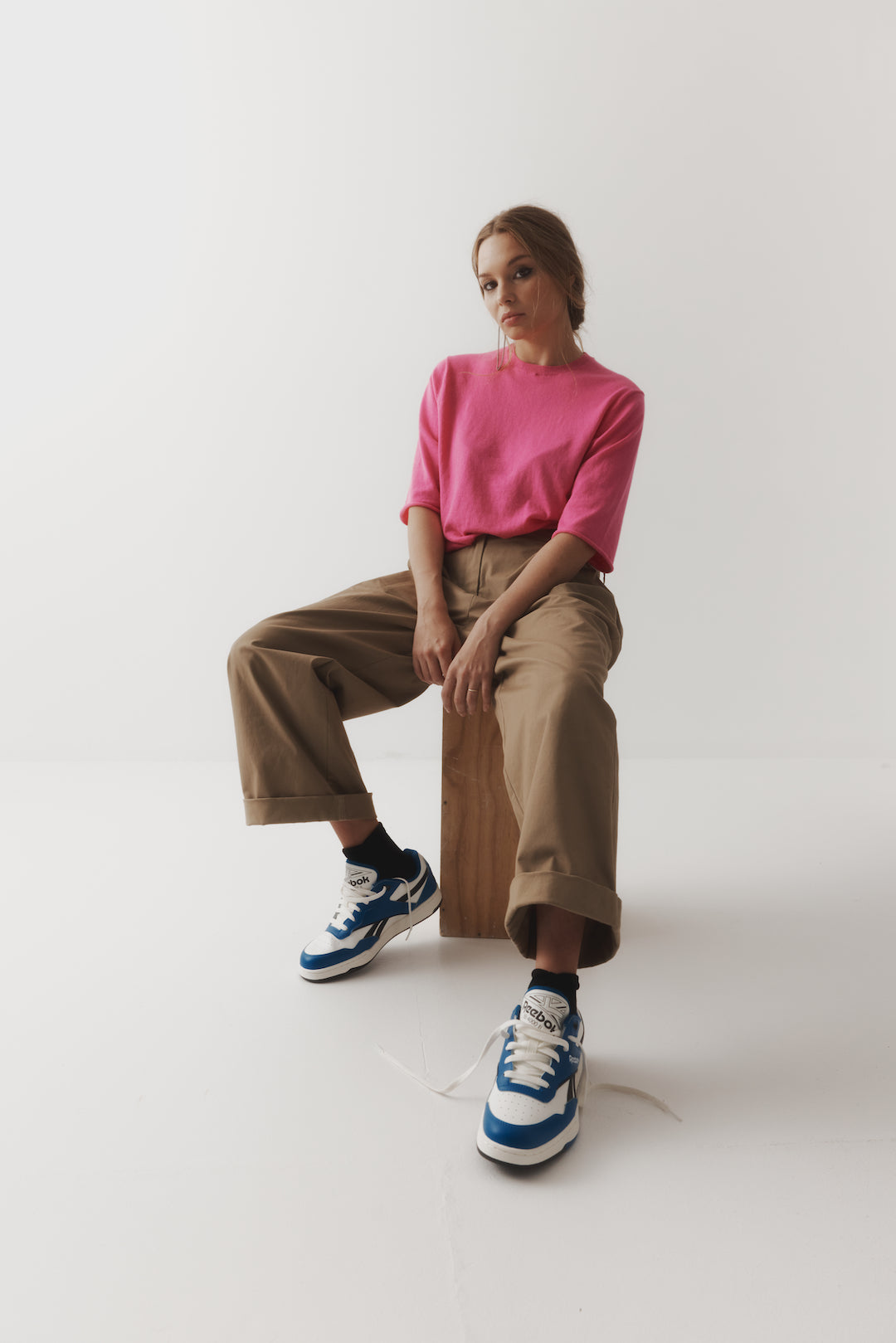 A female model sits on a wooden block in a pink t.shirt with baggy brown cuffed hem pants and sneakers against an ivory backdrop