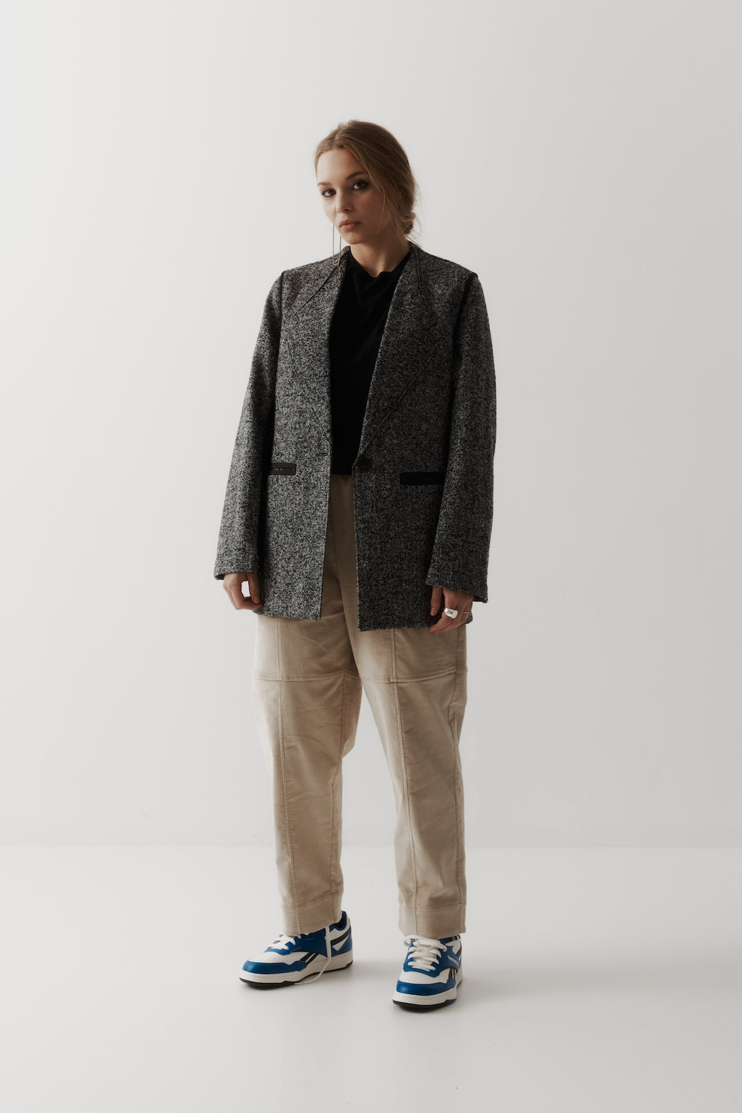 Female model wearing a a textured black and white speckled blazer with black pocket detailing, worn with a black t.shirt and bone coloured corduroy tapered trousers with sneakers. Standing against a plain ivory background