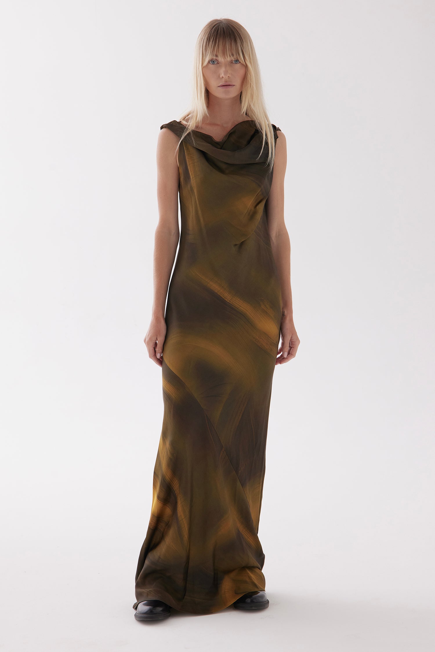 A female model with long straight hair, wears the Overture in Amazon print - a golden and black toned tie dye style print, An off shoulder, one shoulder style or cowl neck style, cut on the bias and full length dress. Featuring beautiful draping through the chest and body.