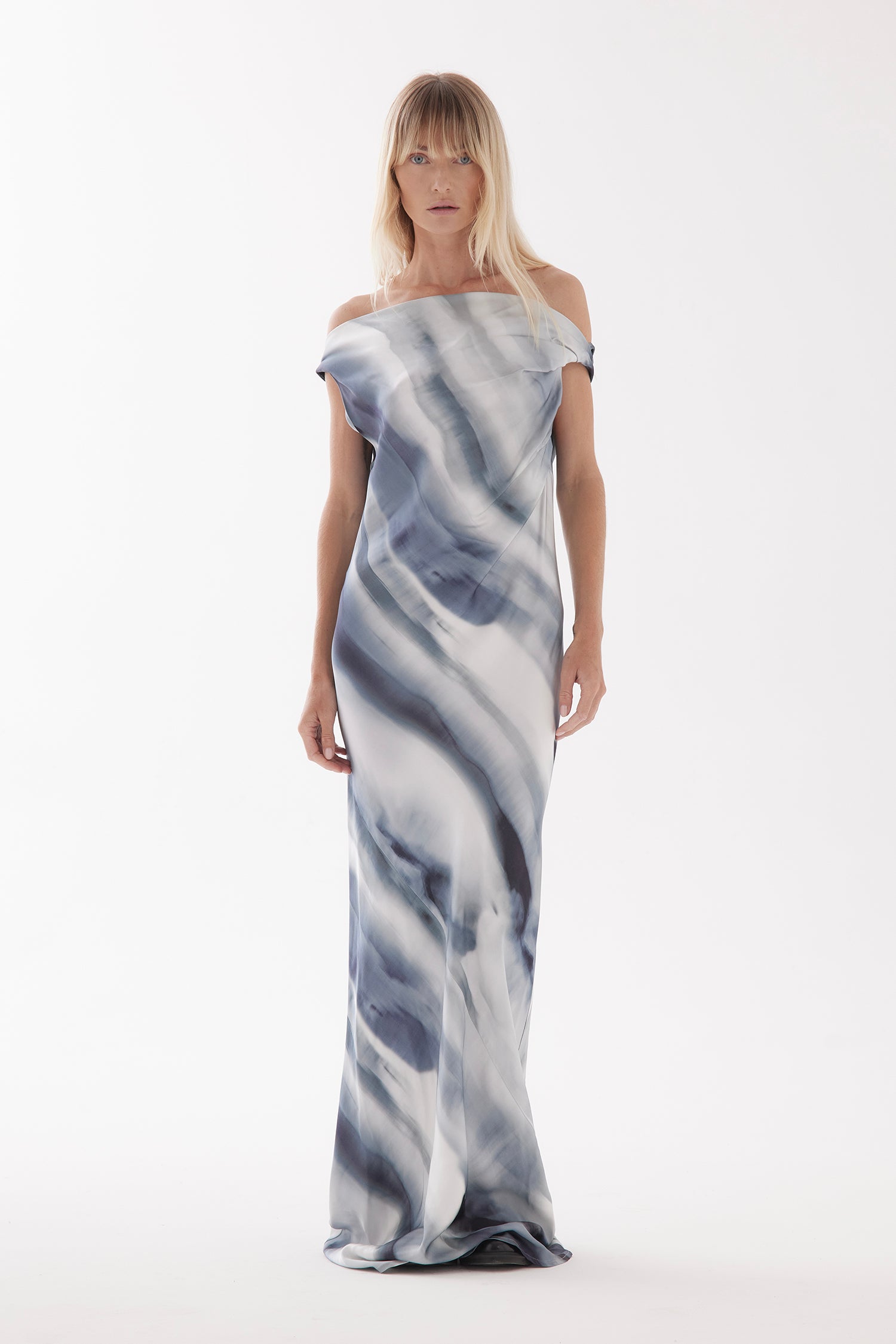 A female model with long straight hair, wears the Overture in Arctic print - a light to dark blue and greys toned tie dye style print. An off shoulder, one shoulder style or cowl neck style, cut on the bias and full length dress. Featuring beautiful draping through the chest and body.