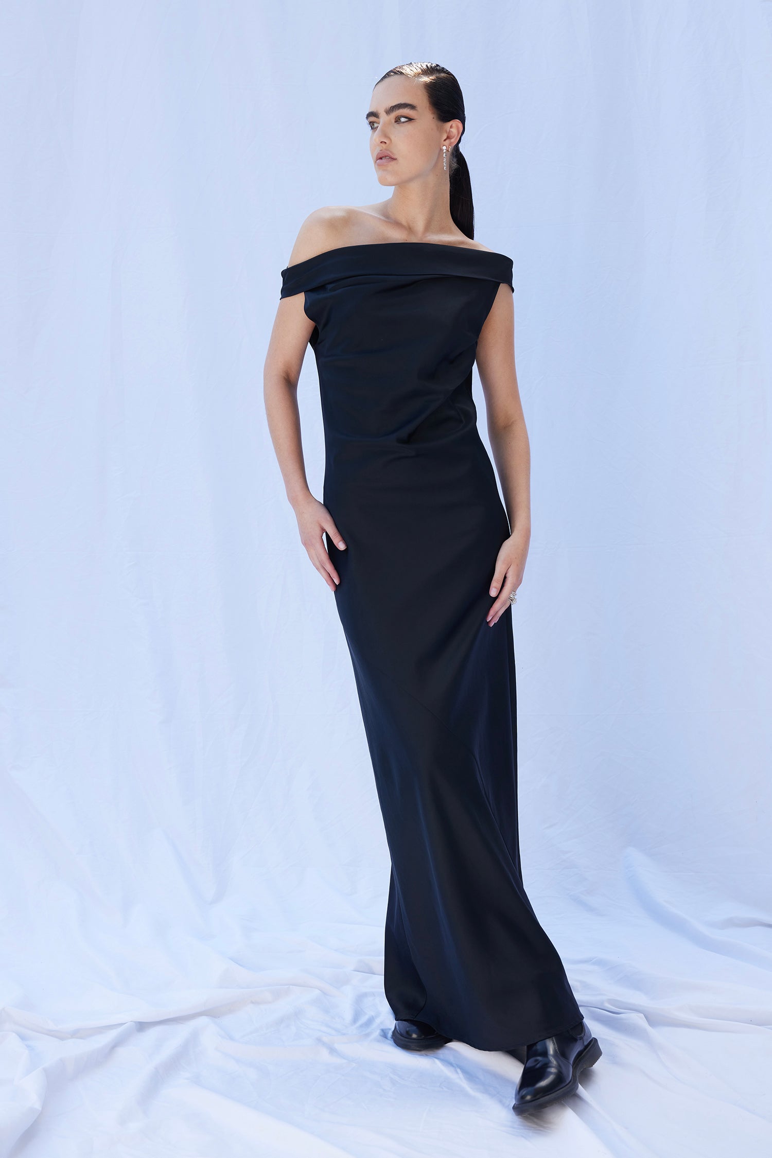 A female model with low slick ponytail wears the Overture in Black, an off shoulder or one shoulder style, cut on the bias and full length. Featuring beautiful draping through the chest and body.