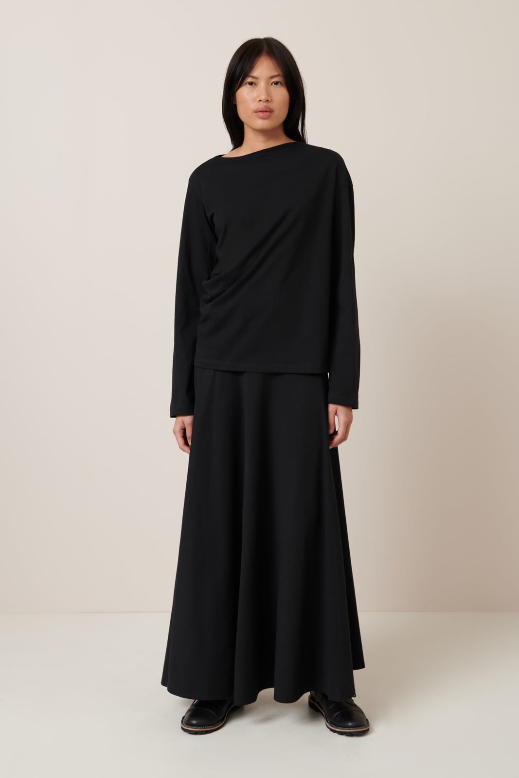 A slender female model wears a 100% organic cotton black top with a minimalist design, and twisted seams that create draped features through the torso a relaxed fit and long sleeves. Worn here with a a-line black cotton jersey full length skirt and black lace up shoes