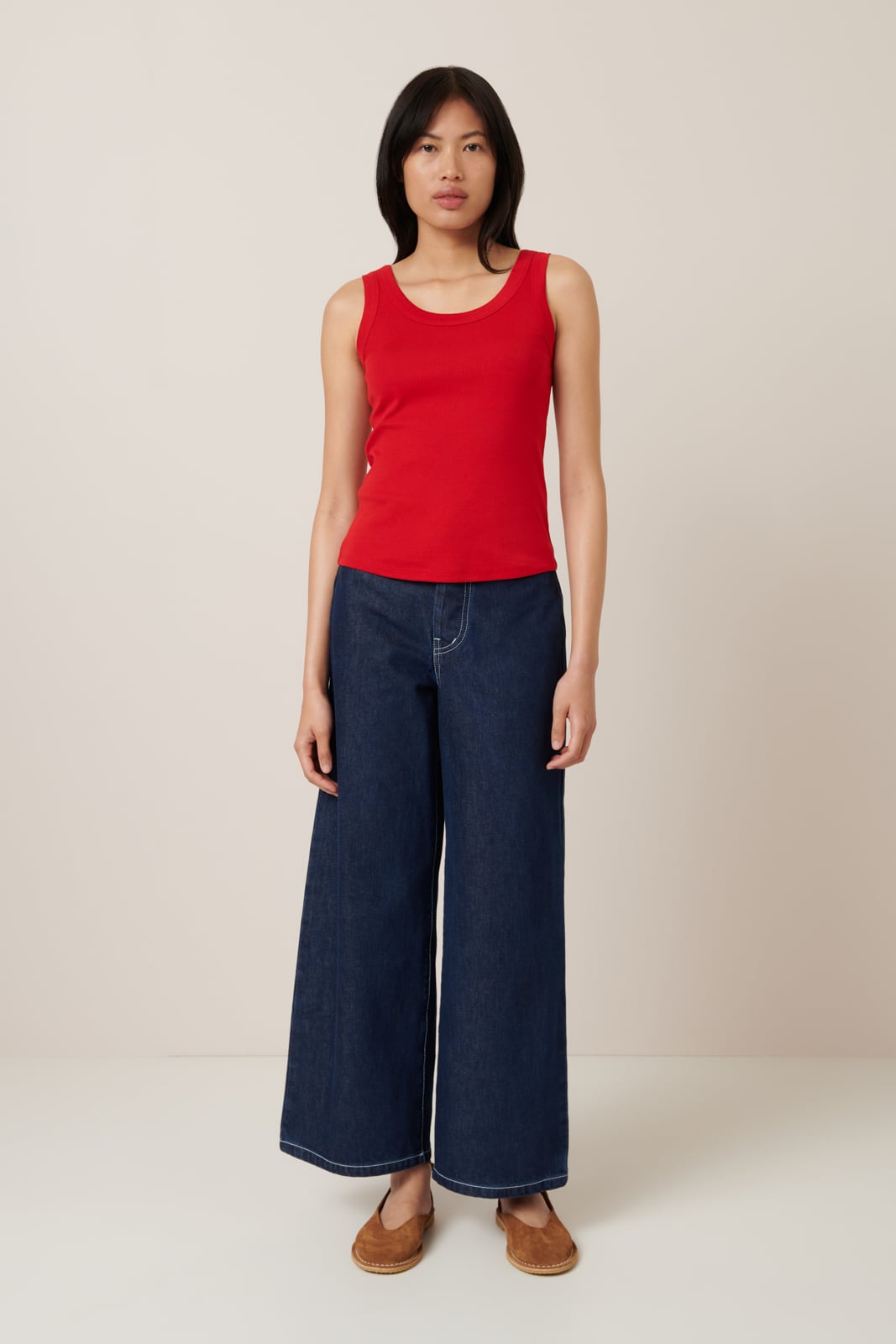 A female model wears a classic red singlet with a simple, sleeveless design, ideal for layering or wearing solo.Worn here with wide leg dark blue jeans and tan ballet flats