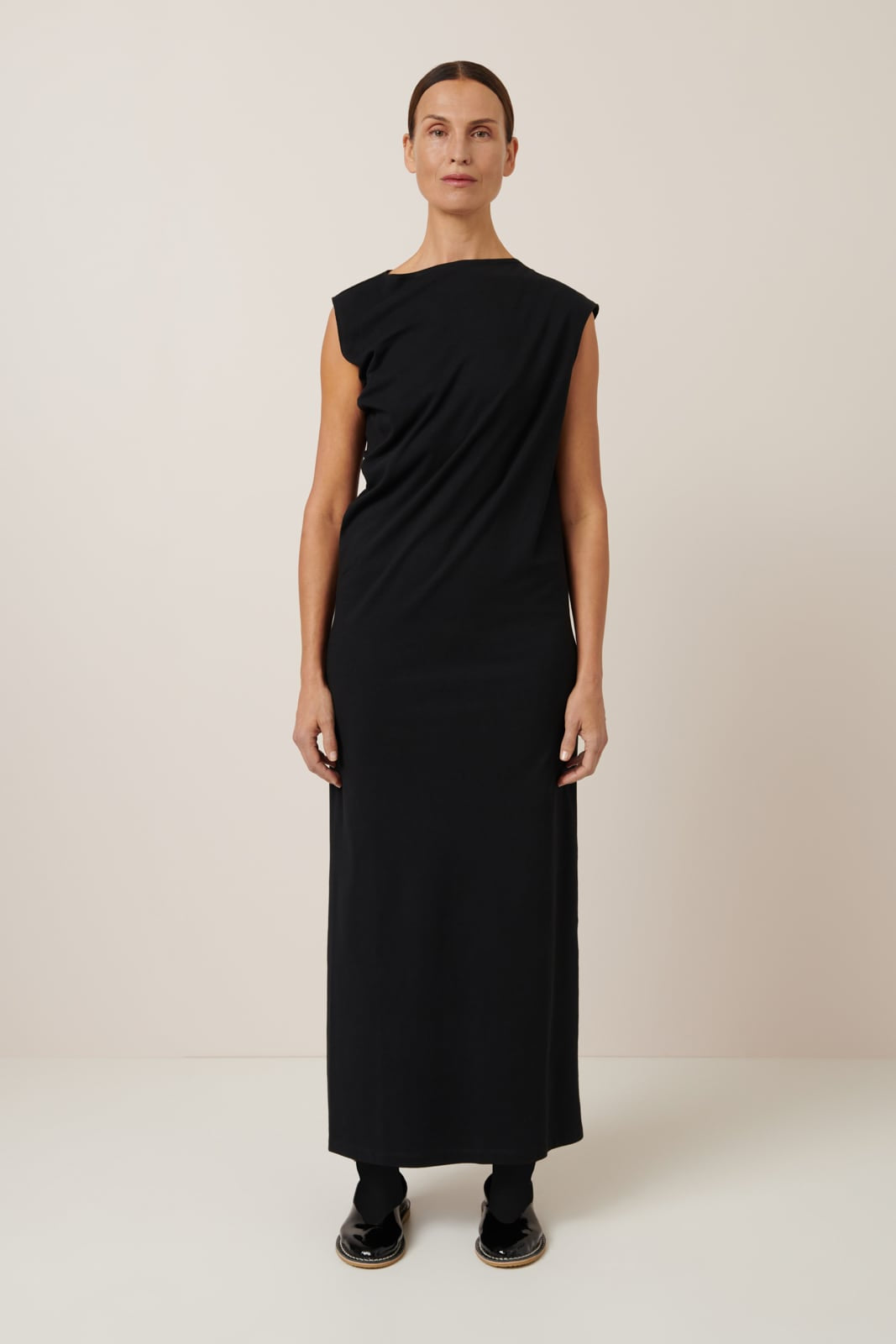 A slender female model wears a full length 100% organic cotton dress, that has an asymmetric neckline, sleeveless and draping through the torso worn with slicked back hair and flat lace up shoes