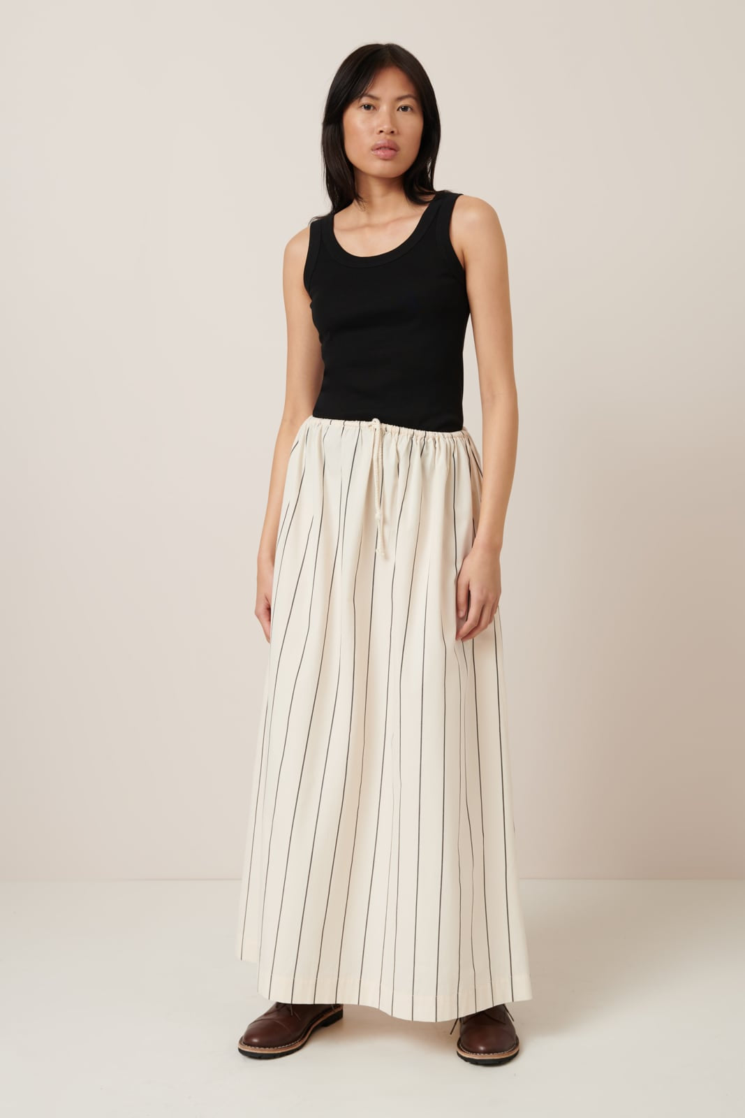 A female model with dark hair wears a greige-colored pinstripe skirt with an elastic waist with draw string, it is full length, straight yet roomy silhouette and worn with a classic black singlet top and brown lace up shoes. 