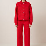 A slender female model with slicked back hair wears a bold red jacket with a structured fit,much like a classic denim jacket featuring silver buttons and 3 pleats in the torso on each side of the buttons. Worn here with matching full length wide leg pants and black lace up shoes 