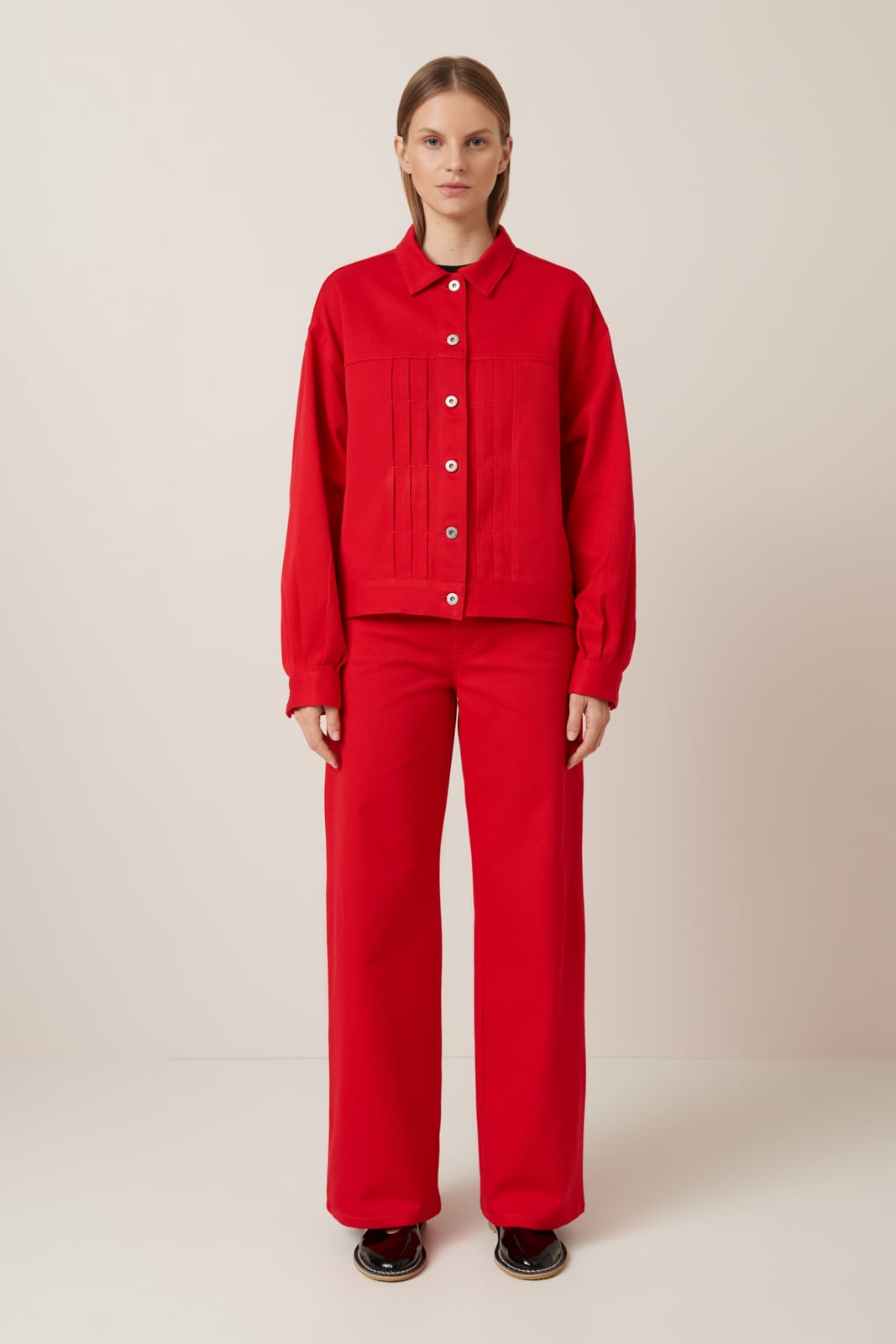 A slender female model with slicked back hair wears a bold red jacket with a structured fit,much like a classic denim jacket featuring silver buttons and 3 pleats in the torso on each side of the buttons. Worn here with matching full length wide leg pants and black lace up shoes 