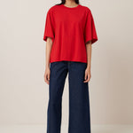 A female model with shoulder length dark hair wears an elbow length, crew neck, oversized fit red t-shirt, it is worn with wide leg dark blue fred short-sleeved t-shirt. Worn here with wide leg dark blue jeans and grey sandals with black socks