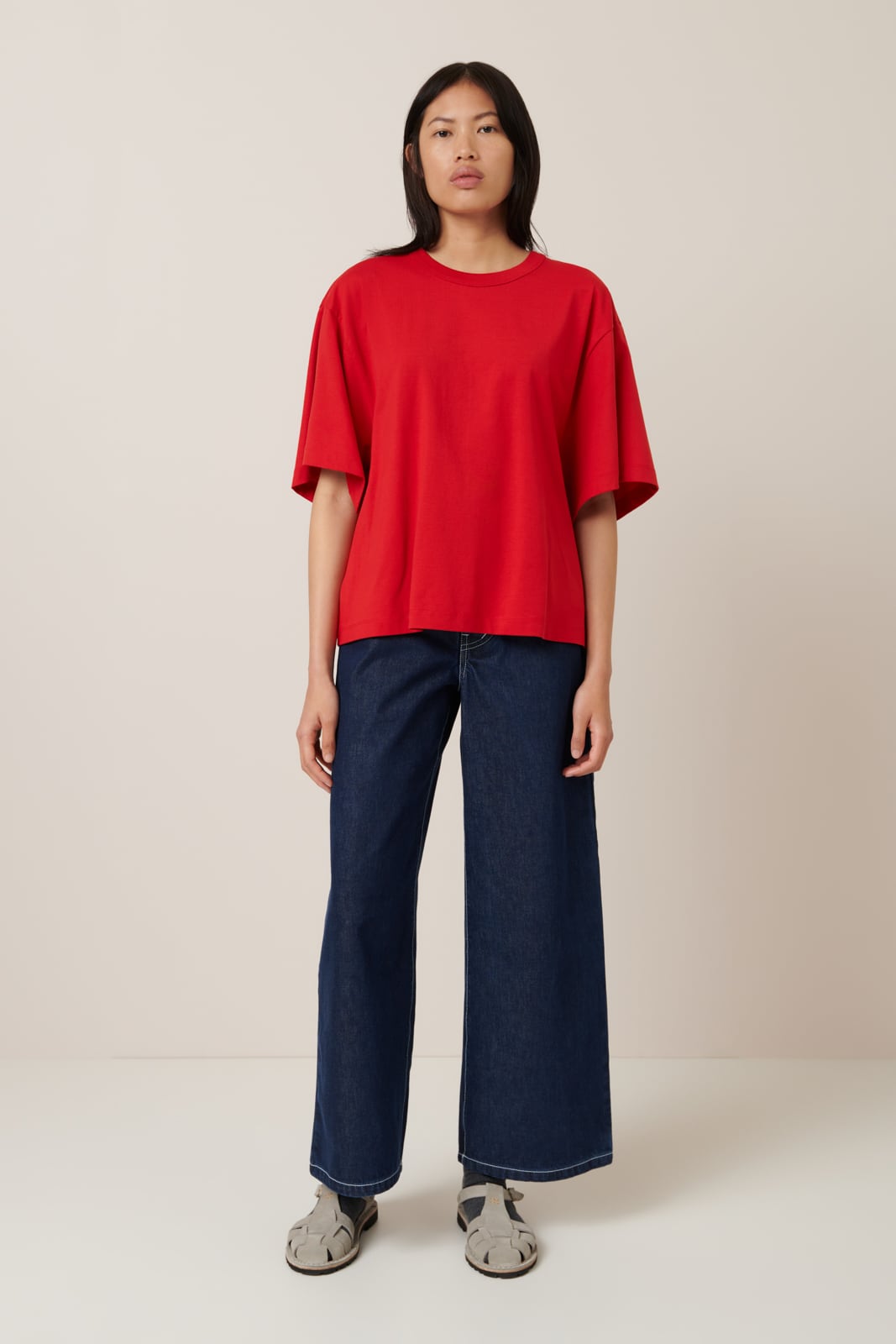 A female model with shoulder length dark hair wears an elbow length, crew neck, oversized fit red t-shirt, it is worn with wide leg dark blue fred short-sleeved t-shirt. Worn here with wide leg dark blue jeans and grey sandals with black socks