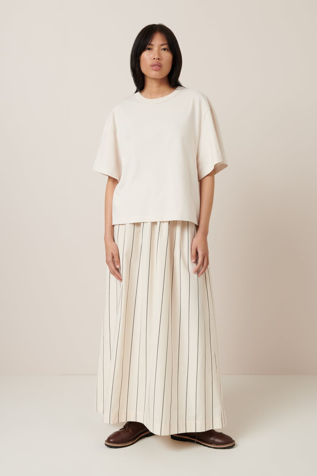 A female model with shoulder length dark hair wears a light-colored short-sleeved t-shirt with an oversized fit and elbow length sleeves made from 100% organic cotton. Worn with a straight full cream and grey pinstripe skirt and flat brown lace up shoes.