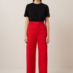 A female model with long slicked back hair wear a pair of vibrant red jeans with a straight wide leg cut and full length hem, they have a high waist and are worn with a classic black t.shirt tucked in and black flat shoes.