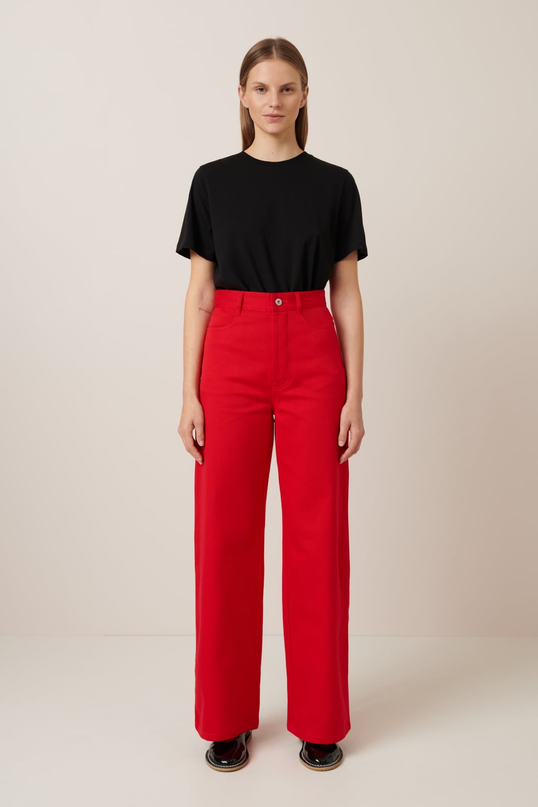 A female model with long slicked back hair wear a pair of vibrant red jeans with a straight wide leg cut and full length hem, they have a high waist and are worn with a classic black t.shirt tucked in and black flat shoes.