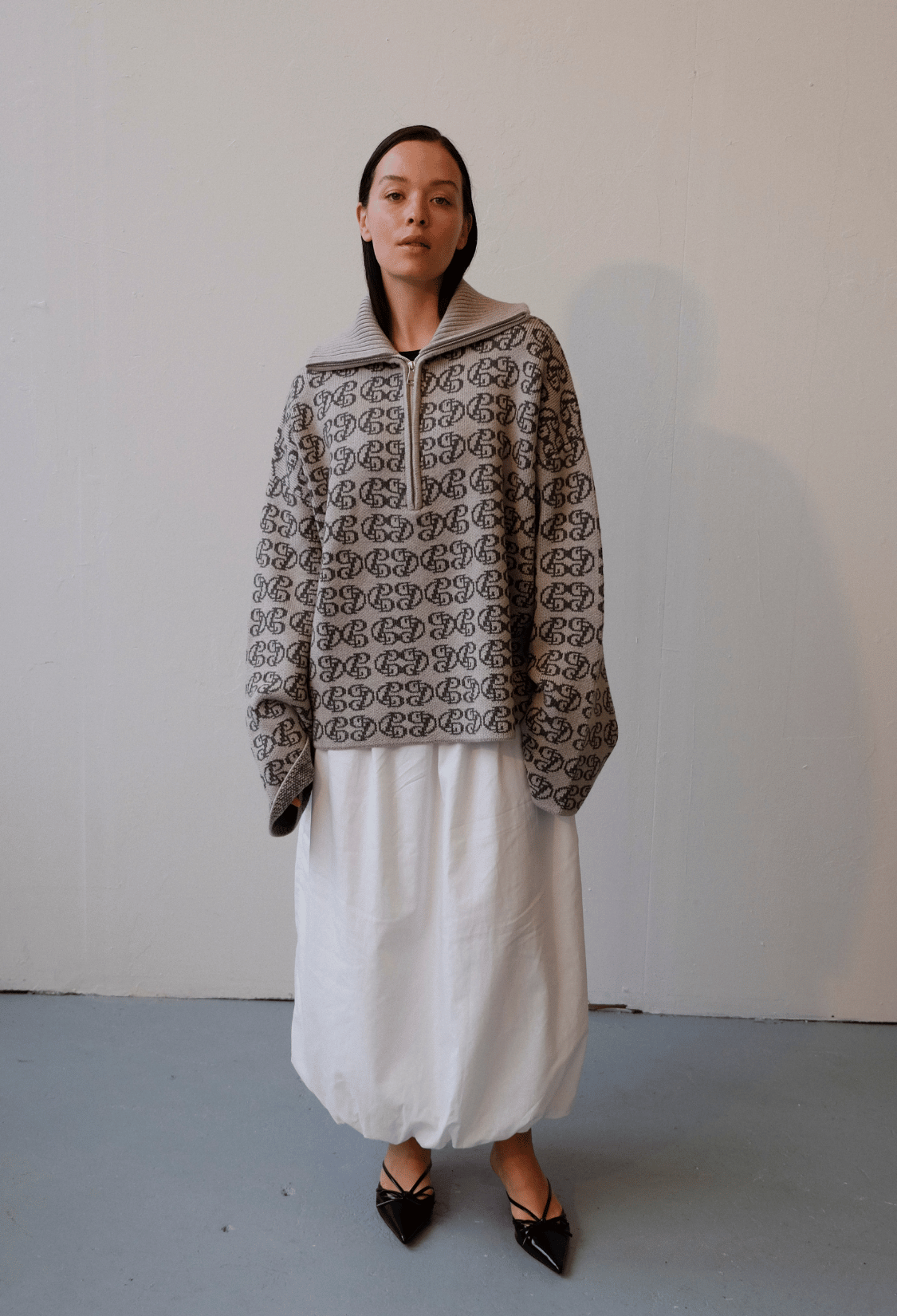 Female model wearing 1/4 Zip Chapman knit, zipped up with collar folded down, and monogram CPD logo repeated all over, in tonal neutral colours. Paired with white cotton bubble skirt and black pointy leather shoes