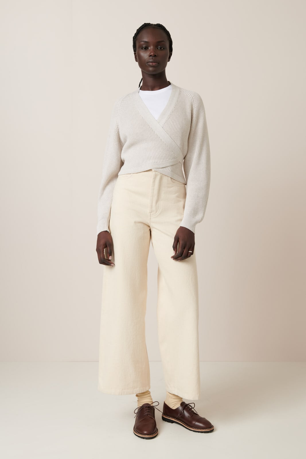 A female model wears a soft oat melange wrap-style cardigan with a  100% organic cotton ribbed knit texture, featuring a deep V-neck and long sleeves. Styled over a white crew neck t.shirt and wide-leg ecru denim wide leg jeans and low lace up brown shoes with beige socks.